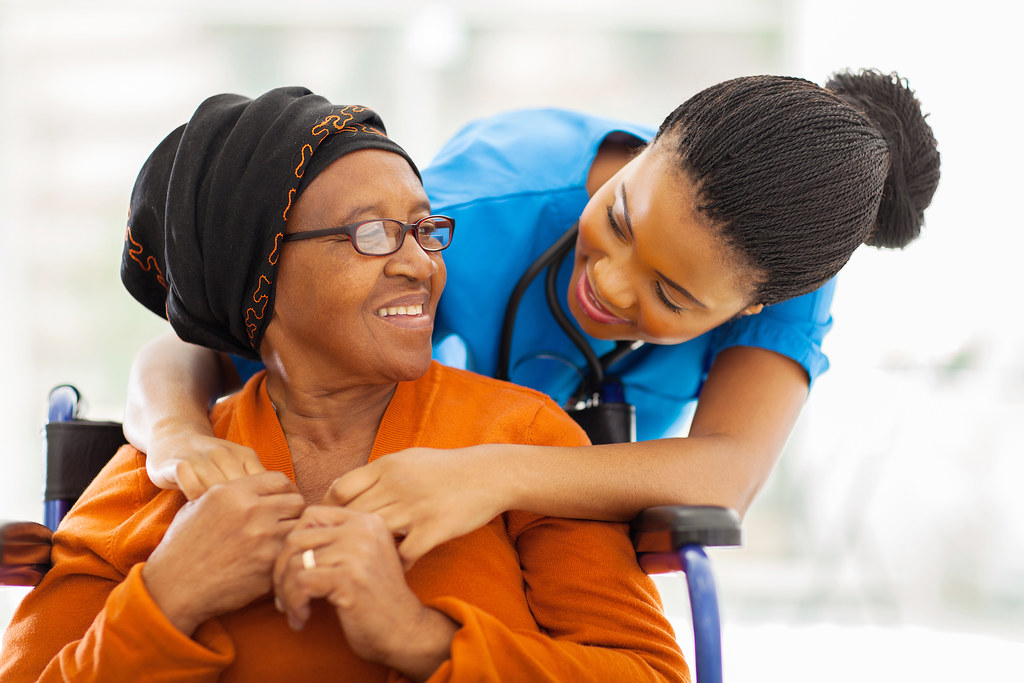 A caregiver in blue scrubs smiles as she leans over the shoulder of her client, an older woman in an orange sweater seated in a wheelchair