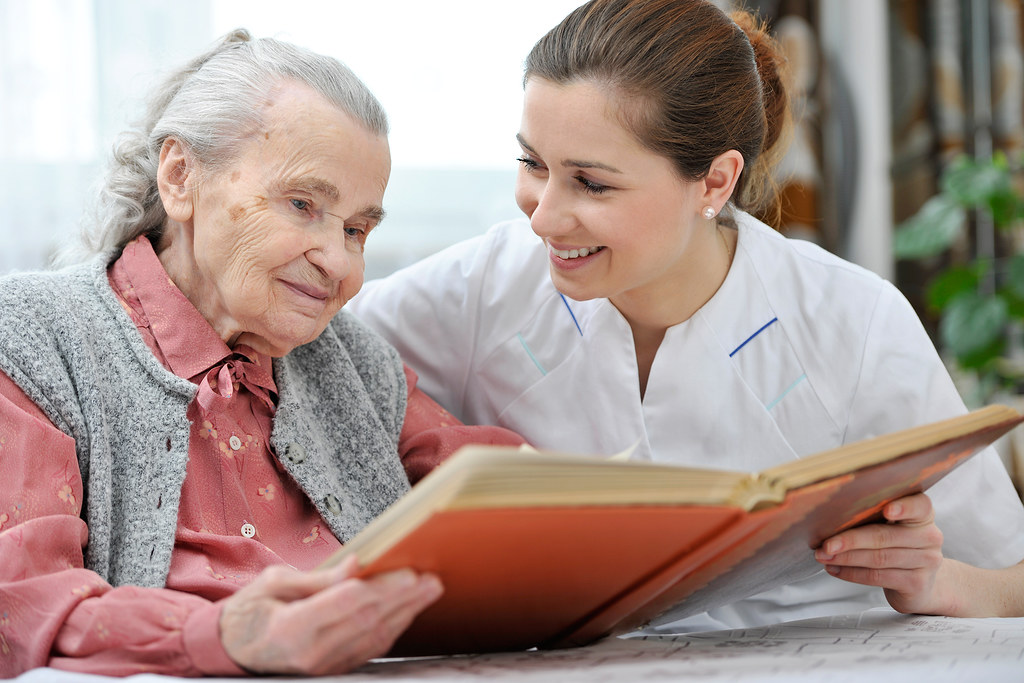 A caregiver in a white uniform reads a book together with her elderly client at the client's home